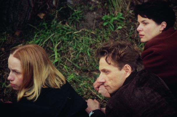 Drei Personen liegen nebeneinander auf Gras, zwei mit braunen Jacken, eine mit schwarzer Kleidung, Blick nach rechts