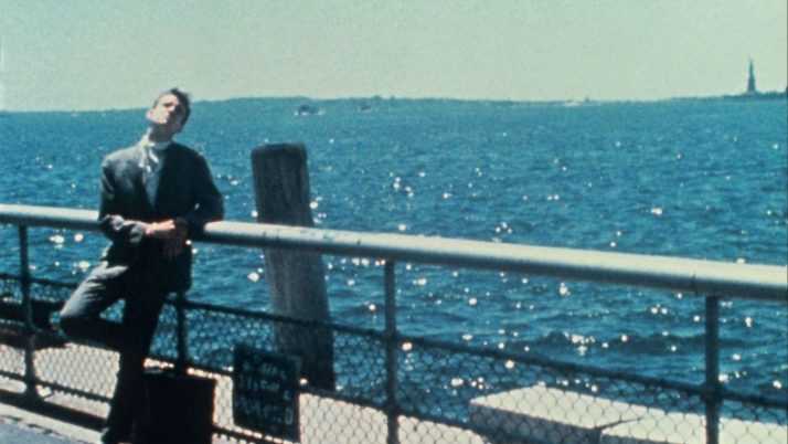 Mann in dunklem Anzug lehnt an einem Geländer mit Blick auf das glitzernde Meer, im Hintergrund die Freiheitsstatue.