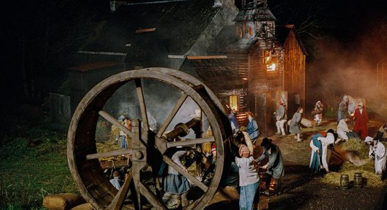 Nachtszene einer historischen Ernteszene mit Menschen in traditioneller Kleidung vor einem großen Holzgebäude und einem hölzernen Wasserrad.