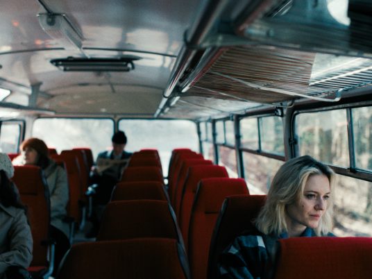 Innenraum eines Busses mit roten Sitzen, mehrere Fahrgäste sitzen verteilt im Hintergrund, eine Frau im Vordergrund blickt aus dem Fenster.