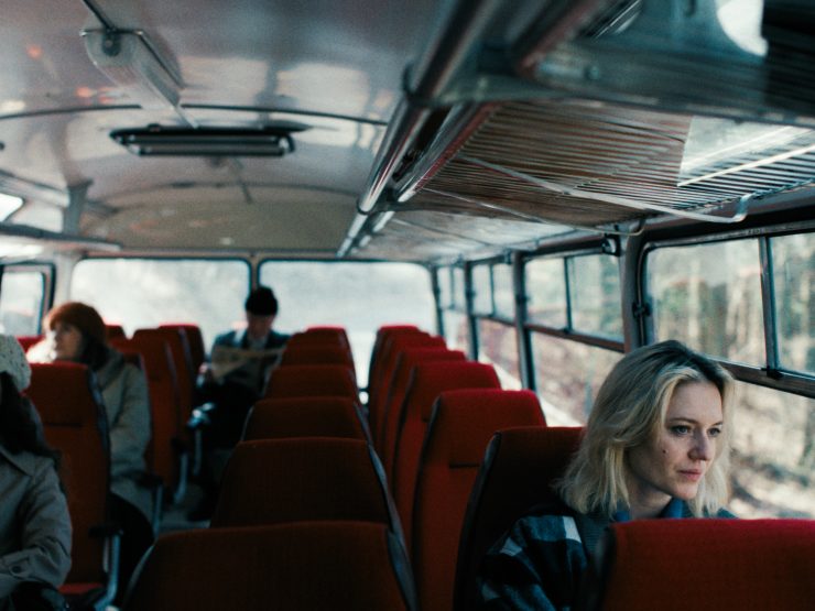 Innenraum eines Busses mit roten Sitzen, mehrere Fahrgäste sitzen verteilt im Hintergrund, eine Frau im Vordergrund blickt aus dem Fenster.