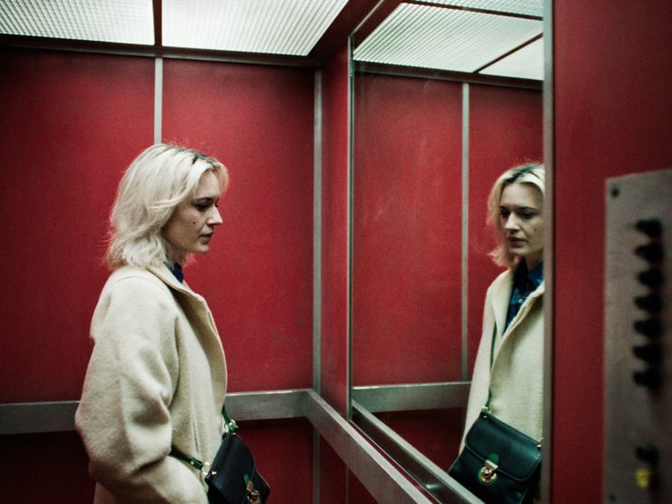 Eine Frau mit hellblondem Haar und beigem Mantel steht in einem roten Fahrstuhl und blickt nachdenklich in den Spiegel.