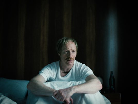 Ein Mann sitzt nachdenklich aufgerichtet in einem Bett, dunkler Hintergrund mit Holzvertäfelung