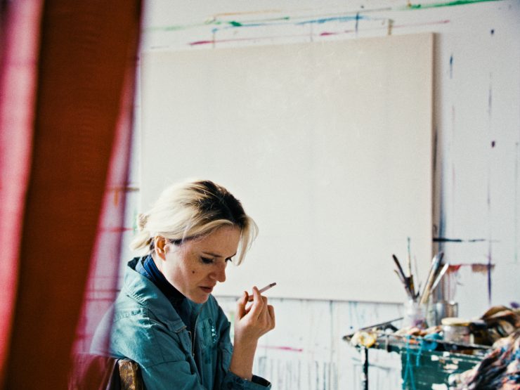 Eine Frau in einem blauem Jeanshemd sitzt in einem Atelier und hält eine Zigarette, hinter ihr ist eine leere Leinwand an der Wand.