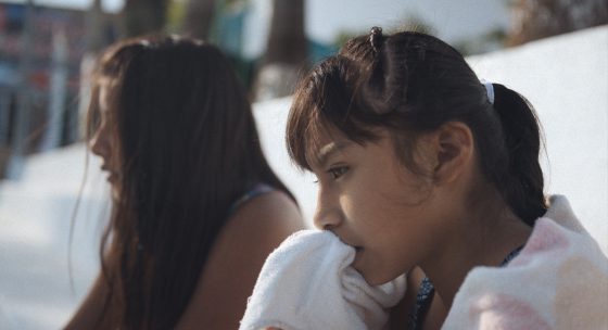 Zwei Mädchen sitzen nebeneinander im Freien; ein Mädchen im Vordergrund hält sich ein Handtuch vor den Mund, beide wirken nachdenklich.