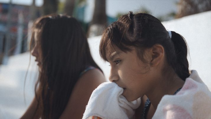 Zwei Mädchen sitzen nebeneinander im Freien; ein Mädchen im Vordergrund hält sich ein Handtuch vor den Mund, beide wirken nachdenklich.