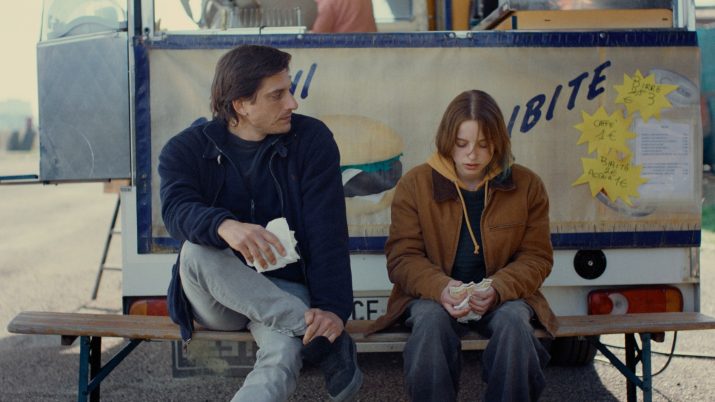 Filmstill "Paternal Leave": Eine weibliche junge Figur und eine männliche ältere sitzen vor einem Imbissstand und unterhalten sich.