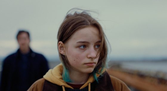 Filmstill "Paternal Leave": Weibliche Figur blickt zur Seite, im Hintergrund eine verschwommene Gestalt - herbstliche Umgebung am Meer.