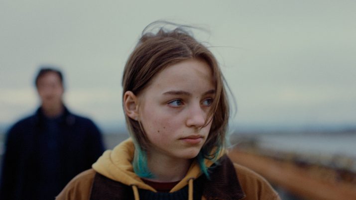 Filmstill "Paternal Leave": Weibliche Figur blickt zur Seite, im Hintergrund eine verschwommene Gestalt - herbstliche Umgebung am Meer.