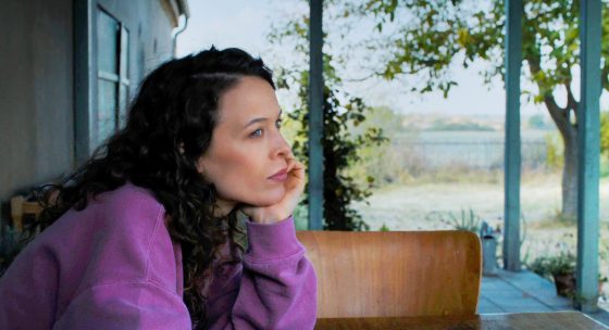 Filmstill aus 'MiroirsNo3': Eine Frau starrt nachdenklich in die Ferne - ländliche Ungembung mit einem See und Haus