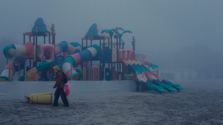 Ein Mensch mit Kapuzenjacke geht an einem verlassenen, bunten Spielplatz mit Rutschen und Klettergerüsten vorbei, umgeben von Nebel.