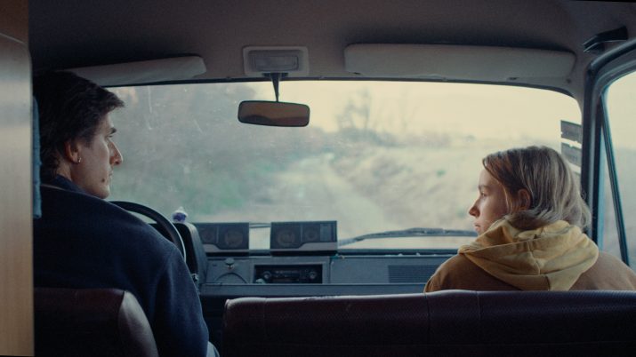 Zwei Personen sitzen vorne in einem Auto, Blick durch die Windschutzscheibe auf eine unbefestigte Straße und eine bewaldete Landschaft.