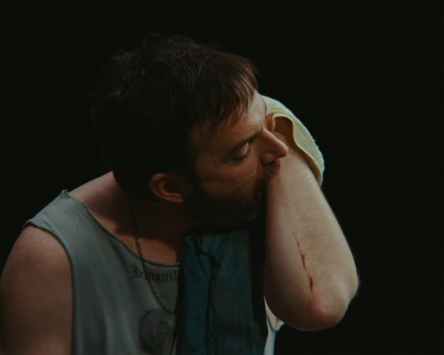 Eine Person vor schwarzem Hintergrund berührt mit dem Mund den eigenen Unterarm.