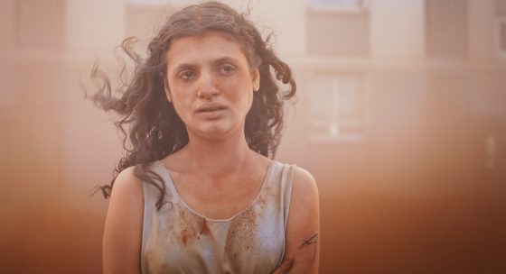 Eine junge Frau mit traurigen Blick ist umgeben und teils bedeckt von beigem Sand.