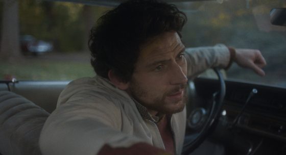 Filmstill: Ein Mann sitzt am Lenkrad im Auto, die Kamera blickt auf ihn vom Rücksitz aus. Er stützt einen Arm auf das Lenkrad, den anderen Arm auf die Rücklehne.