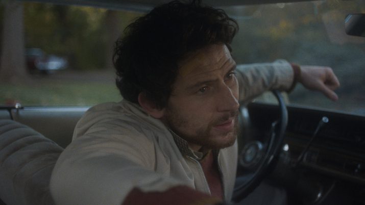Filmstill: Ein Mann sitzt am Lenkrad im Auto, die Kamera blickt auf ihn vom Rücksitz aus. Er stützt einen Arm auf das Lenkrad, den anderen Arm auf die Rücklehne.