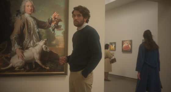 Filmstill: Ein Mann steht vor einem Gemälde in einer Ausstellung. Eine Hand in der Hosentasche, in der anderen hält er einen Schlüssel.