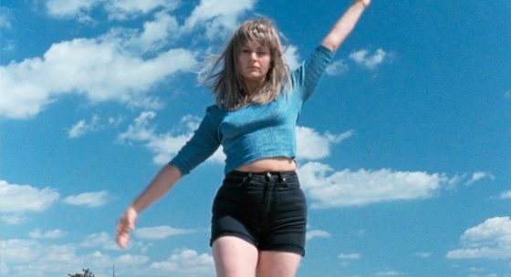 Frau mit blauem, langärmligem Oberteil und schwarzen Shorts springt mit erhobenen Armen vor blauem Himmel mit Wolken