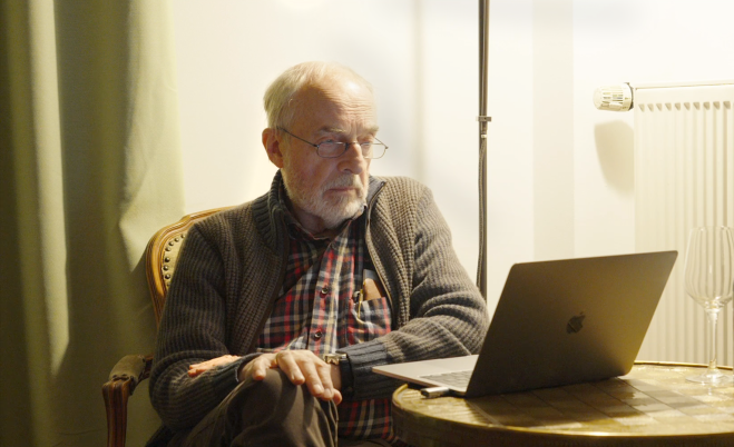 Älterer Mann mit grauem Strickpullover und kariertem Hemd sitzt auf einem Stuhl neben einem runden Holztisch mit einem geöffneten MacBook und einem leeren Weinglas.