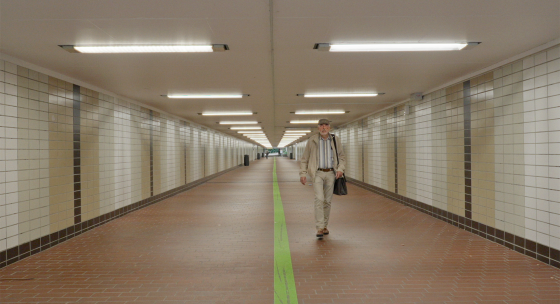Ein Mann in beigem Anzug und gestreiftem Hemd geht in einem langen, beleuchteten Tunnel mit braunem Boden und weißen Kacheln an den Wänden, eine grüne Linie verläuft mittig auf dem Boden.