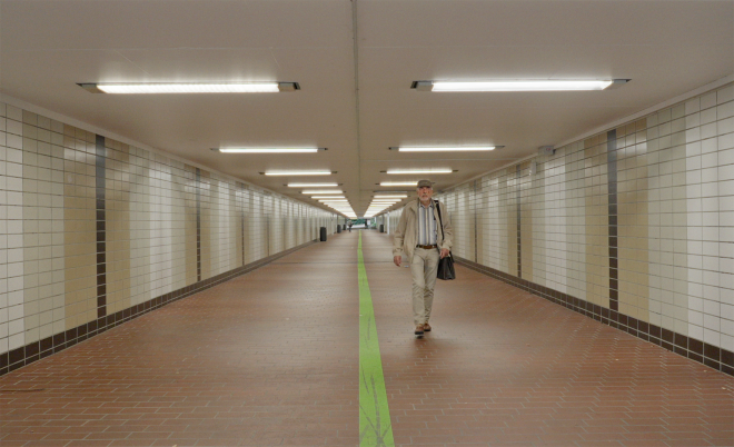 Ein Mann in beigem Anzug und gestreiftem Hemd geht in einem langen, beleuchteten Tunnel mit braunem Boden und weißen Kacheln an den Wänden, eine grüne Linie verläuft mittig auf dem Boden.