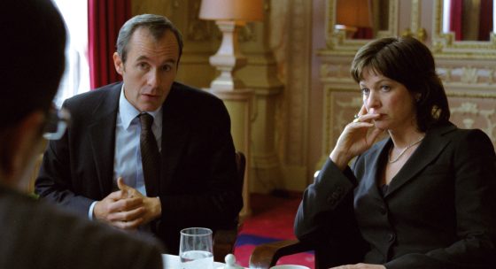 Zwei Personen in formeller Kleidung sitzen an einem Tisch in einem elegant eingerichteten Raum mit rotem Teppich und goldfarbenen Wänden.