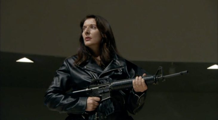 Person mit langen dunklen Haaren trägt schwarze Lederjacke und hält ein Gewehr in beiden Händen.