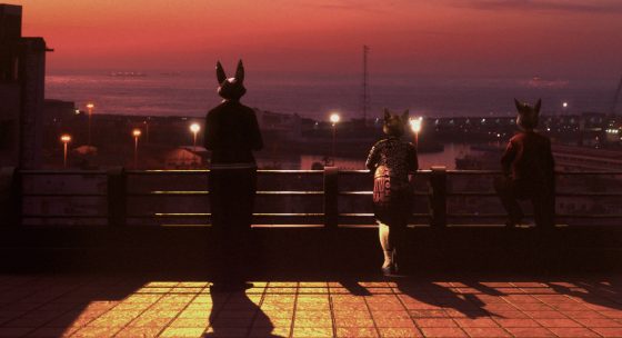 Drei Personen mit Tiermasken stehen auf einer Terrasse und blicken auf eine beleuchtete Stadt bei Sonnenuntergang.