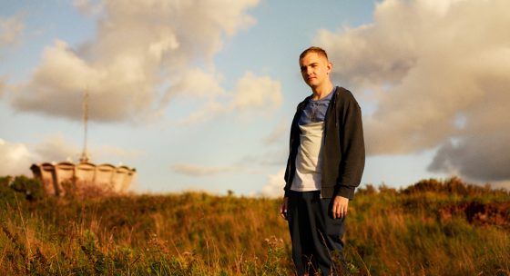 Person steht auf einer Wiese mit langem Gras, trägt schwarze Jacke, weißes T-Shirt und dunkle Hose, im Hintergrund Wolken und ein Bauwerk.