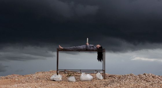 Person liegt auf dem Rücken auf einem Metallgestell, dunkle Wolken am Himmel, felsiger Boden mit weißen Steinen