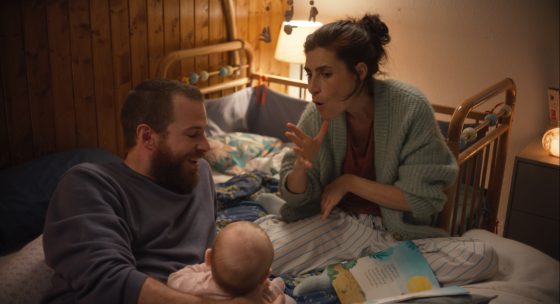 Zwei Erwachsene sitzen auf einem Bett, eine Person hält ein Baby, eine andere gestikuliert mit der Hand. Im Hintergrund Holzvertäfelung und eine Lampe.