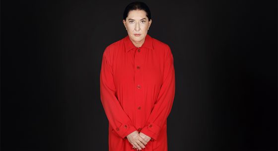 Person steht vor schwarzem Hintergrund, trägt rotes, langärmliges Hemd mit Knöpfen, Hände vor dem Körper verschränkt.