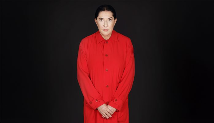 Person steht vor schwarzem Hintergrund, trägt rotes, langärmliges Hemd mit Knöpfen, Hände vor dem Körper verschränkt.