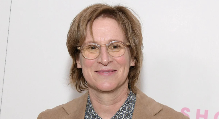 Person mit kurzen, blonden Haaren trägt hellbraunen Blazer und blau-weiß gemustertes Hemd vor weißer Wand mit rosa Schrift.