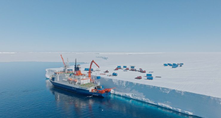 Großes Forschungsschiff mit orangefarbenen Kränen liegt an einer hohen, schneebedeckten Eiswand, mehrere kleine Fahrzeuge und Container auf dem Eis im Hintergrund.