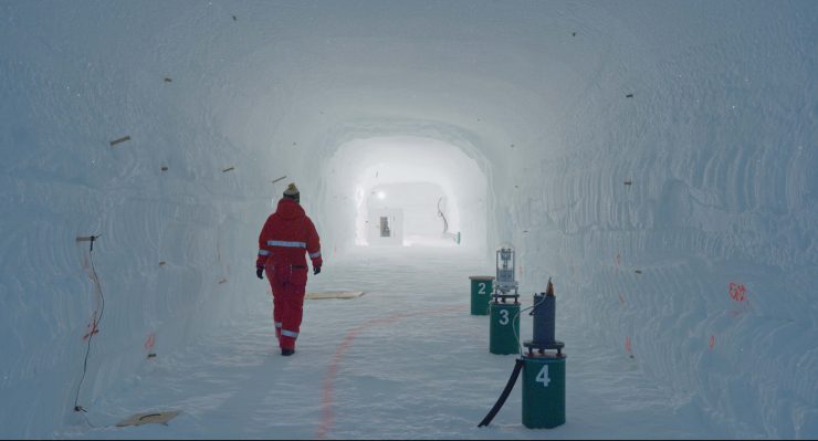Person in rotem Schutzanzug geht durch einen langen, weißen Tunnel mit nummerierten grünen Behältern an der rechten Seite.
