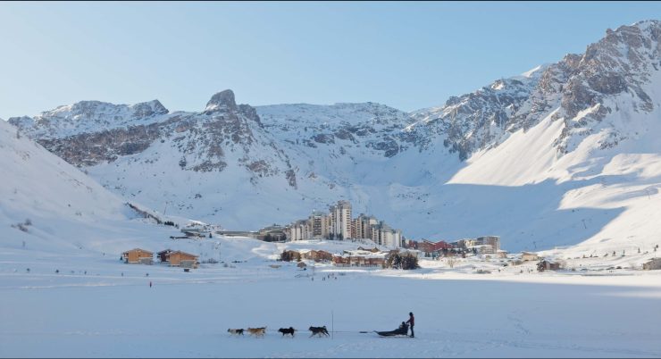 Schneebedeckte Berglandschaft mit einem Dorf am Fuß der Berge und einem Hundeschlitten mit mehreren Hunden im Vordergrund.