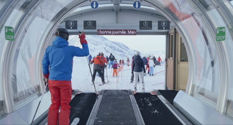 Skifahrer in bunter Winterkleidung steigen aus einer überdachten Förderbandröhre auf eine schneebedeckte Piste mit Bergen im Hintergrund.