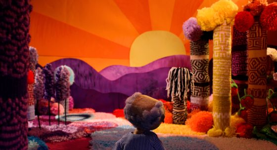 Figur mit braunen Haaren und blau-weiß gestreiftem Hemd blickt auf eine bunte Landschaft aus gestrickten Säulen, Bäumen und Hügeln vor einem orangefarbenen Sonnenuntergang.