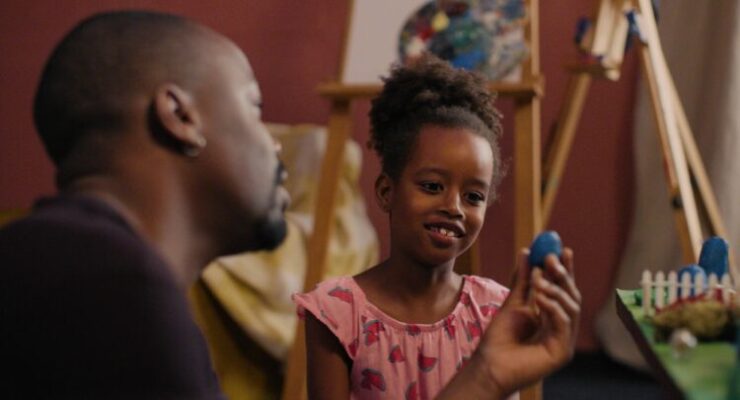 Mwita Mataro zeigt einem Mädchen eine blaue Spielfigur. Das Mädchen lächelt.