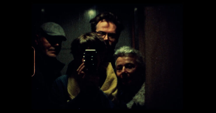 Drei Personen stehen eng beieinander in einem dunklen Raum, eine Person hält eine Kamera vor das Gesicht.