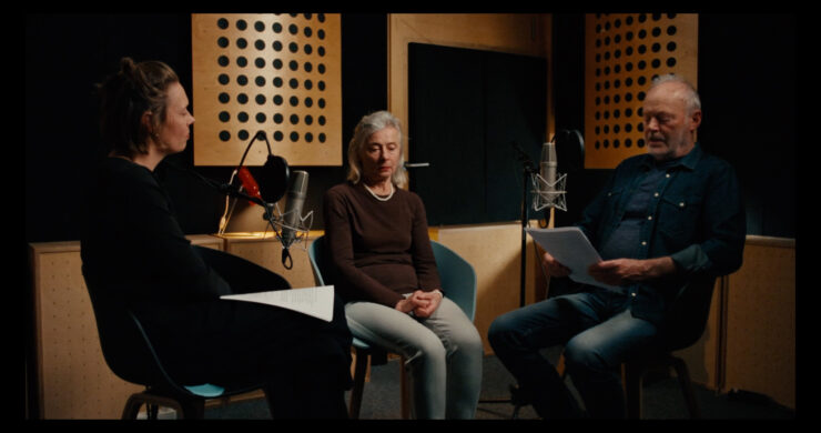 Drei Personen sitzen in einem Tonstudio vor Mikrofonen, zwei halten Papiere in der Hand