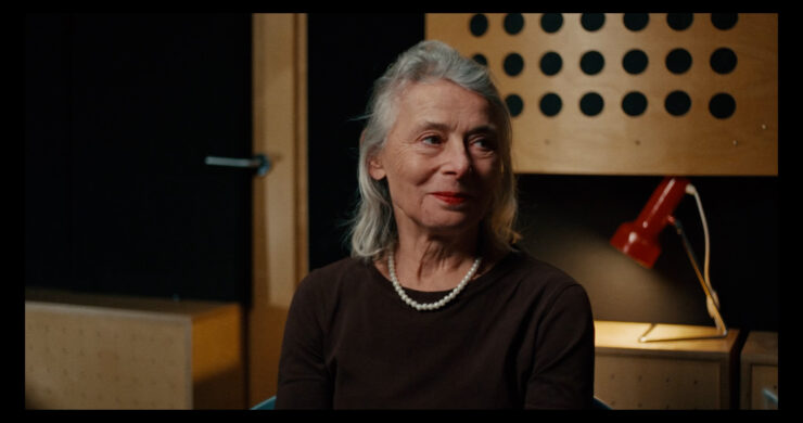 Eine ältere Frau mit grauen Haaren trägt eine Perlenkette und ein dunkles Oberteil vor einer Wand mit Schallabsorberplatten und einer roten Schreibtischlampe.