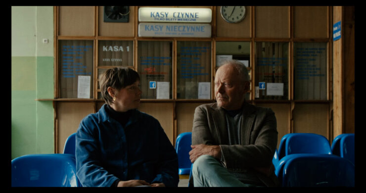 Zwei Personen sitzen nebeneinander auf blauen Stühlen vor einem Schalter mit den Schildern 'KASA 1', 'KASY CZYNNÉ' und 'KASY NIECZYNNE'