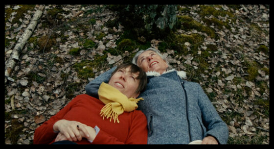 Zwei Personen liegen nebeneinander auf einem mit Laub bedeckten Waldboden, eine trägt einen gelben Schal und rote Kleidung, die andere eine graue Jacke.