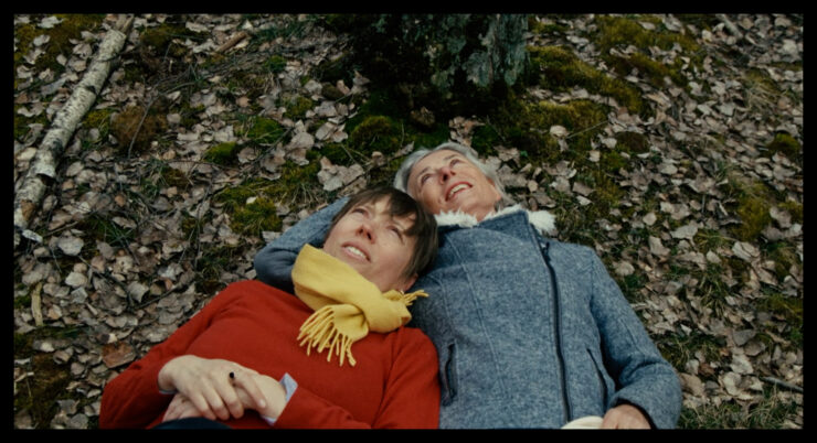 Zwei Personen liegen nebeneinander auf einem mit Laub bedeckten Waldboden, eine trägt einen gelben Schal und rote Kleidung, die andere eine graue Jacke.