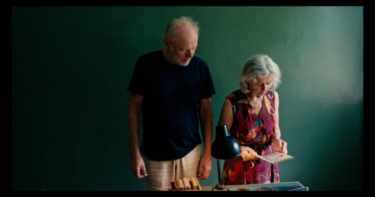 Ein älterer Mann in dunkelblauem T-Shirt und eine ältere Frau in gemustertem Kleid stehen vor einer grünen Wand, die Frau hält ein Buch oder Heft