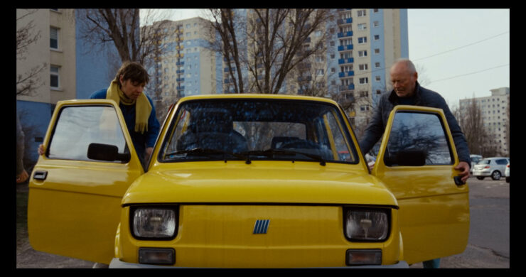 Gelber Kleinwagen mit geöffneten Vordertüren steht vor Hochhäusern, zwei Personen lehnen an den Türen.