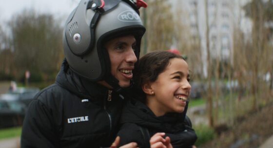 Person mit Helm und schwarzer Jacke hält ein Kind mit schwarzer Jacke auf einem Motorrad oder Roller im Freien fest