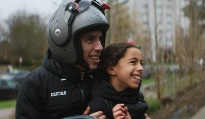 Person mit Helm und schwarzer Jacke hält ein Kind mit schwarzer Jacke auf einem Motorrad oder Roller im Freien fest